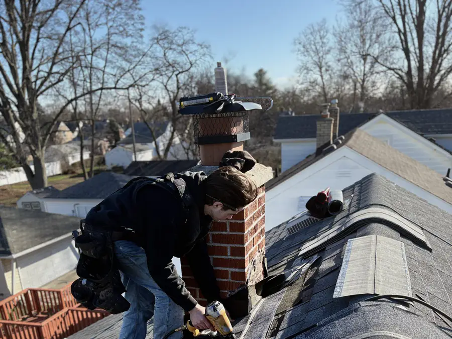 A Joe’s Roofing crew member nailing ridge cap shingles around a brick chimney.