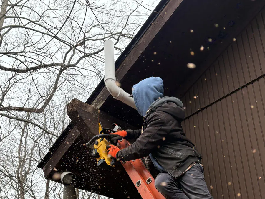 A roofer on a ladder fitting fascia board beside a downspout on a gable end.