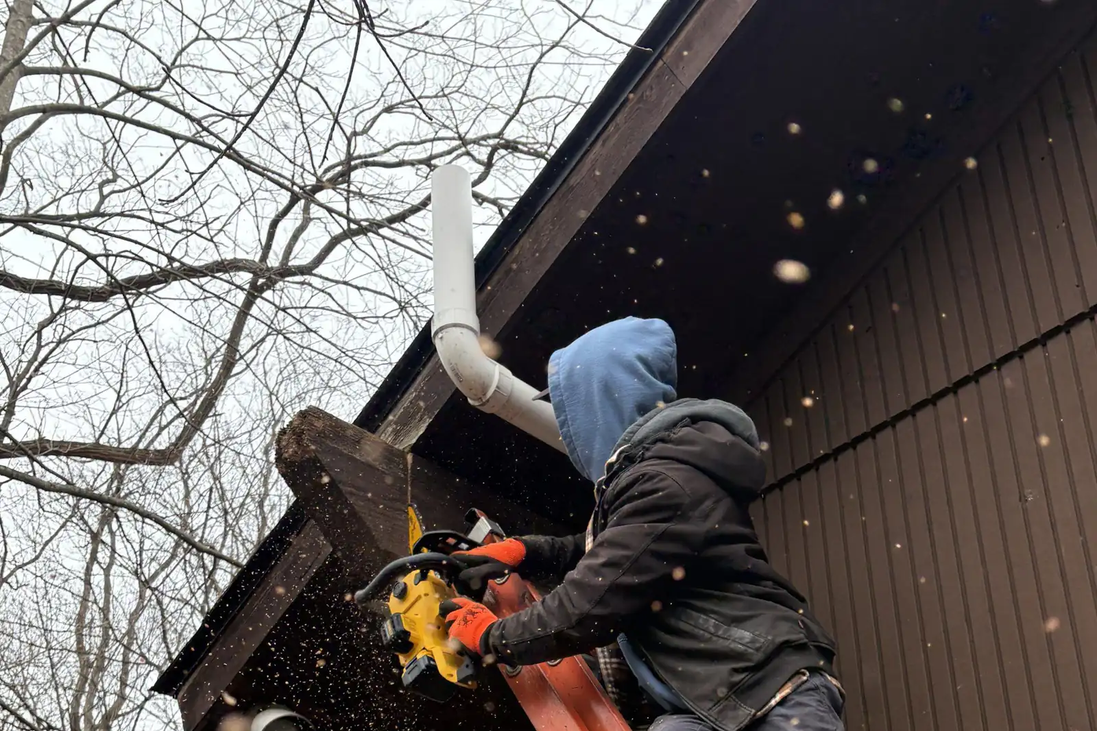 A roofer on a ladder fitting fascia board beside a downspout on a gable end.
