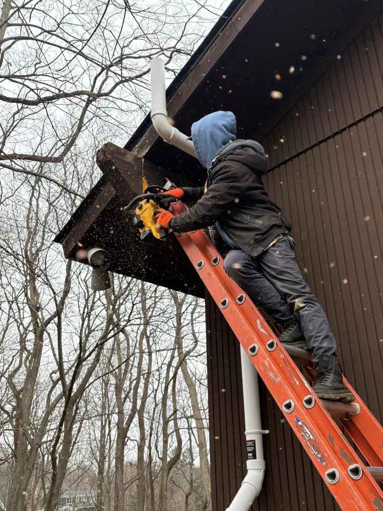 A roofer on a ladder fitting fascia board beside a downspout on a gable end.