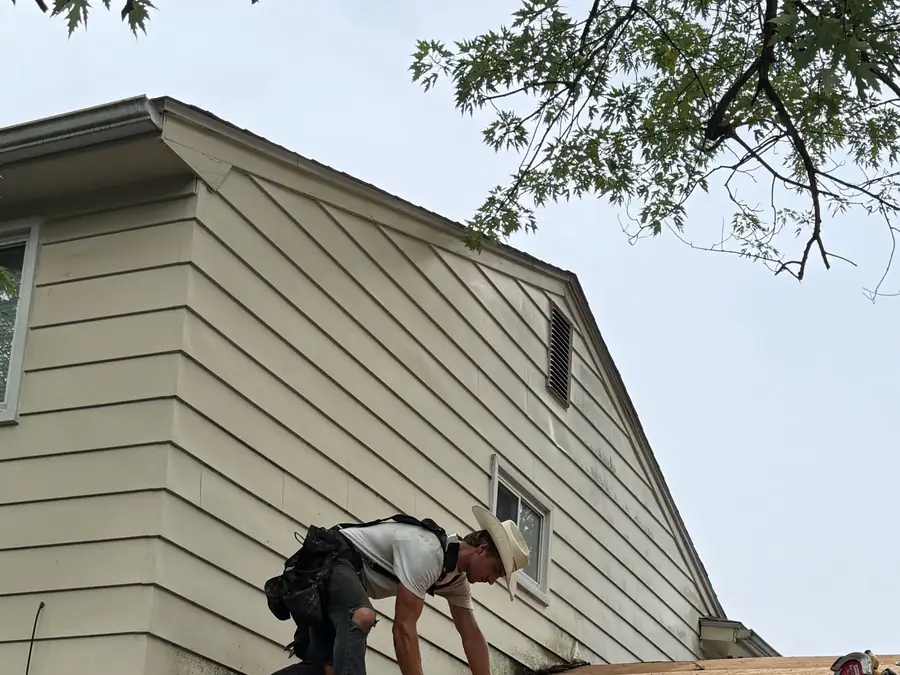 A roofer tearing off old shingles down to the deck on the gable end of a two-storey house.