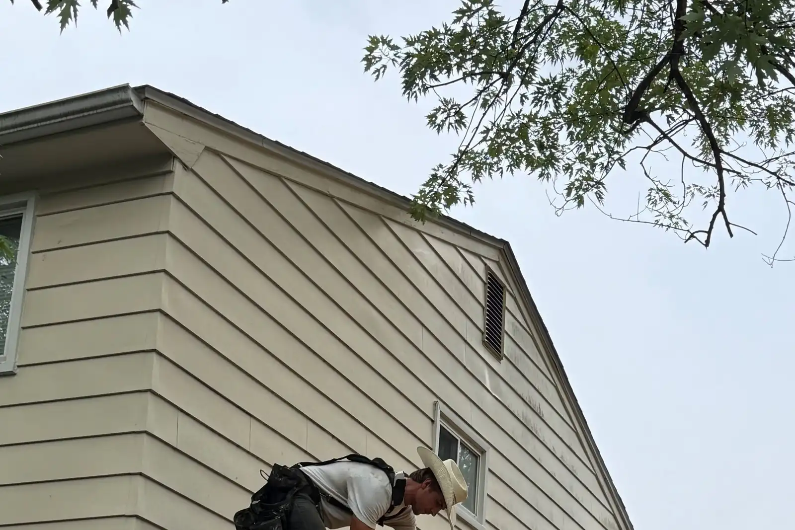 A roofer tearing off old shingles down to the deck on the gable end of a two-storey house.