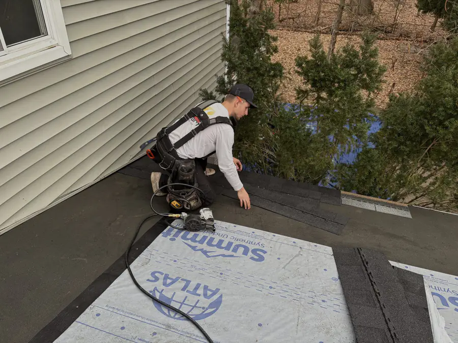 A roofer rolling out synthetic underlayment across a roof deck beside the siding.