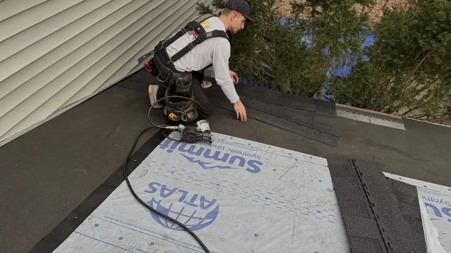 A roofer rolling out synthetic underlayment across a roof deck beside the siding.